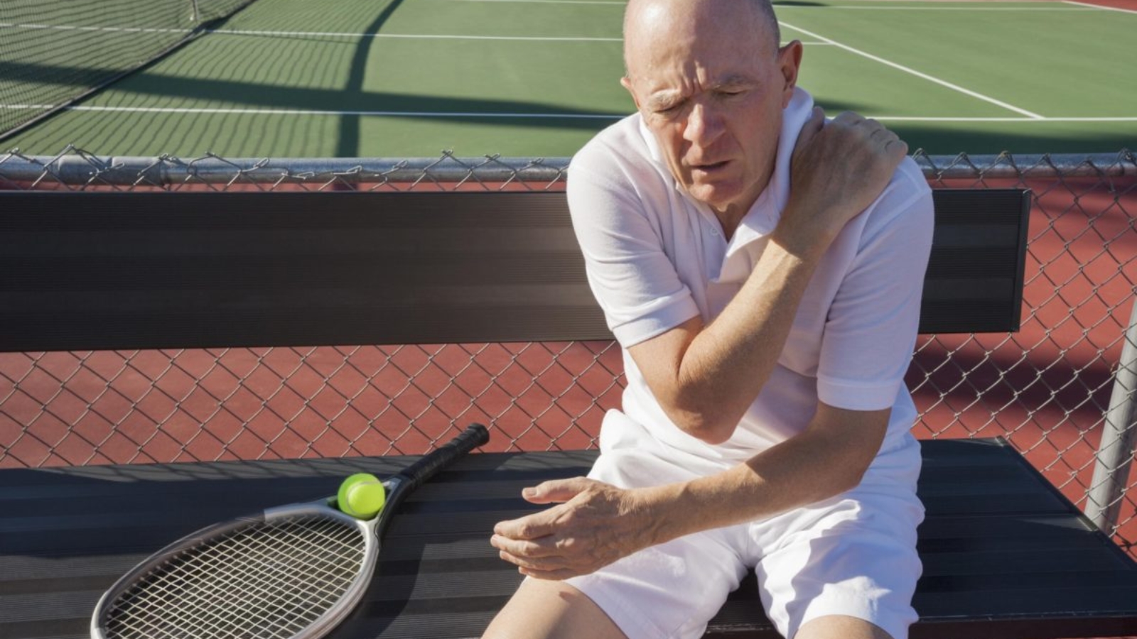 Senior male tennis player with shoulder pain sitting on bench at court